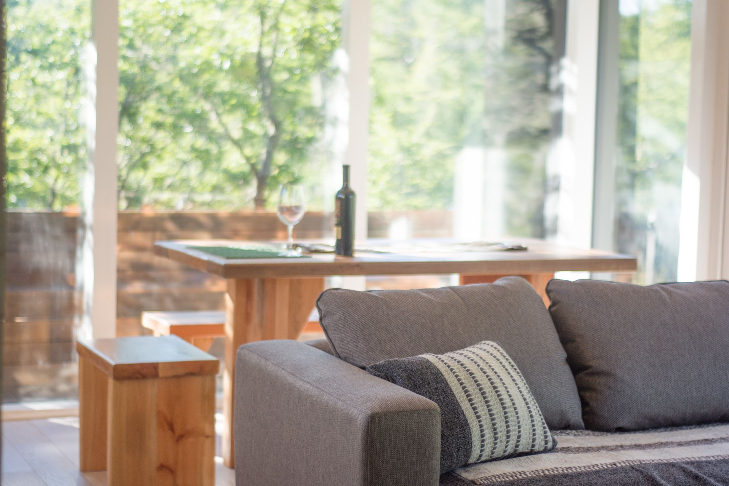 Een moderne woonkamer met een grijze bank, een houten salontafel en een wijnglas op een houten tafel. Grote ramen bieden uitzicht op groen buiten.
