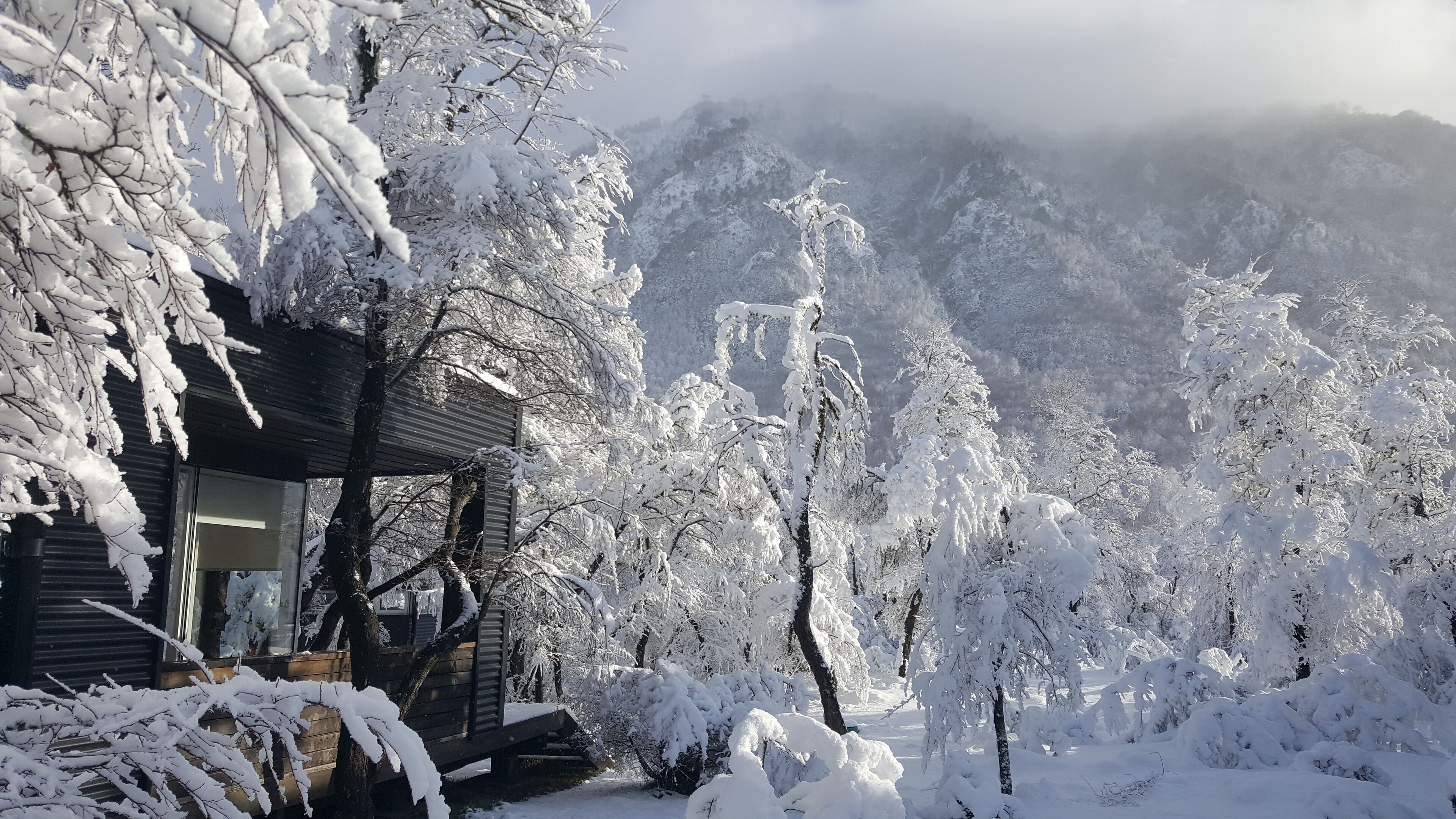 A snow-covered cabin with a wooden deck and a mountain backdrop.