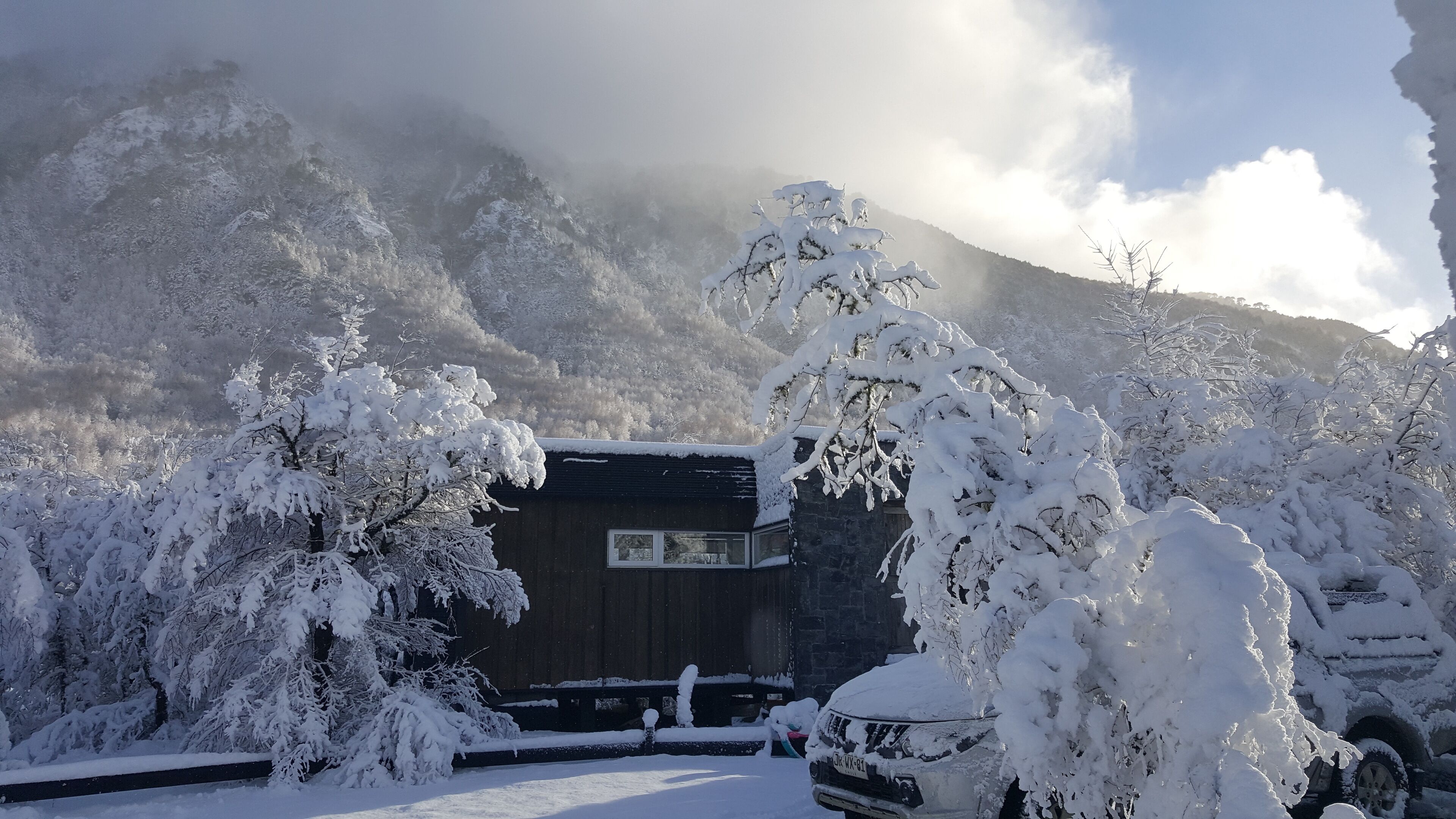 A wooden cabin with a dark roof, surrounded by snow-covered trees and a mountain backdrop.