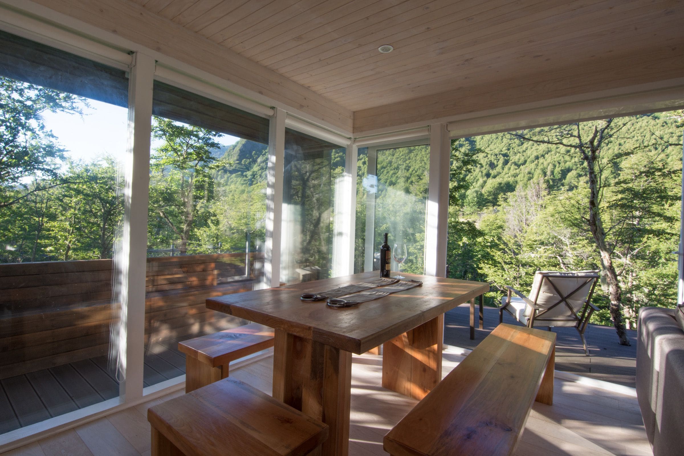 An outdoor wooden deck with a table and benches, surrounded by large glass windows offering a view of a forested area.