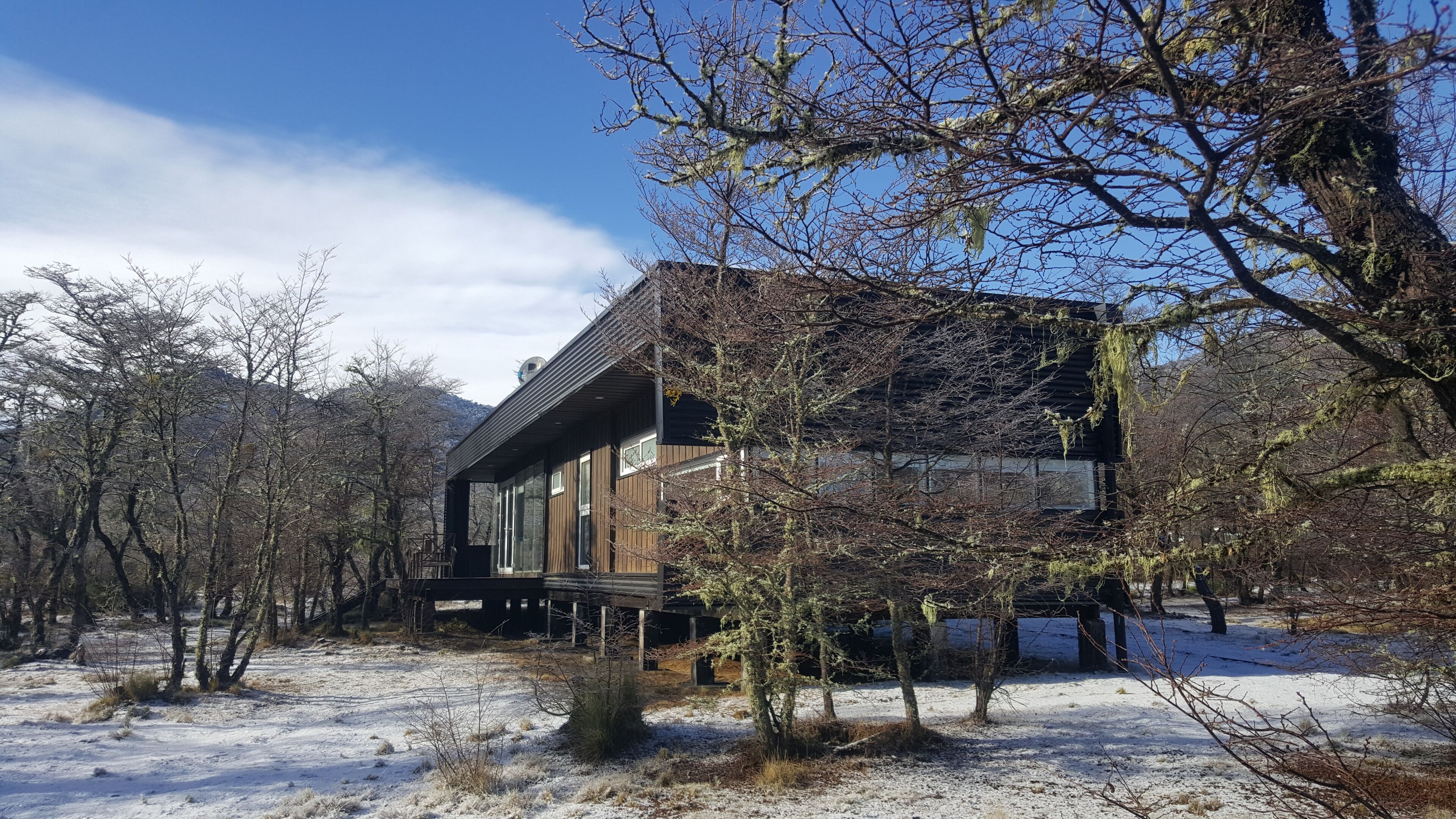 A modern wooden cabin surrounded by snow-covered ground and leafless trees.