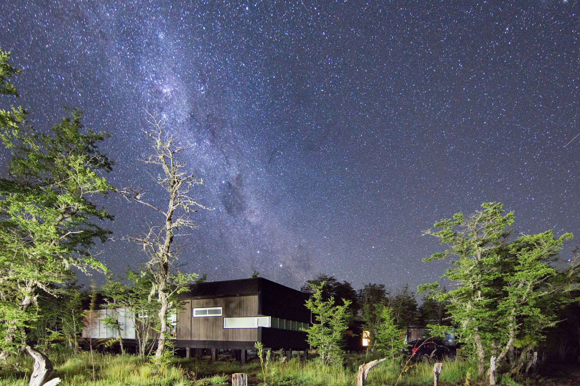 A modern house with a large window, surrounded by trees and a starry night sky.