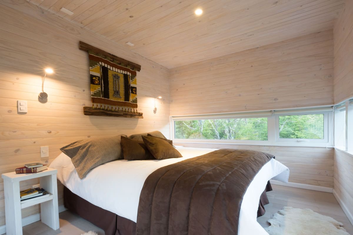 A bedroom with a large bed, a nightstand, a wall-mounted artwork, and a window with a view of trees.