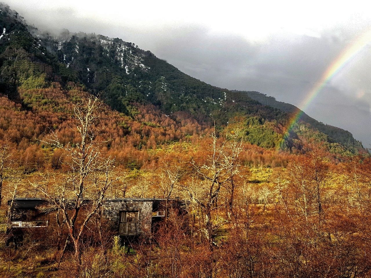 A mountainous landscape with a rainbow, autumn-colored trees, and a rustic stone structure.