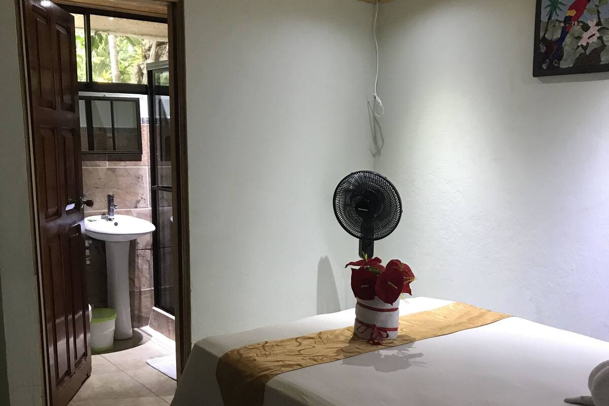 A hotel room with a bed, a bedside fan, a window, and a door leading to a bathroom.