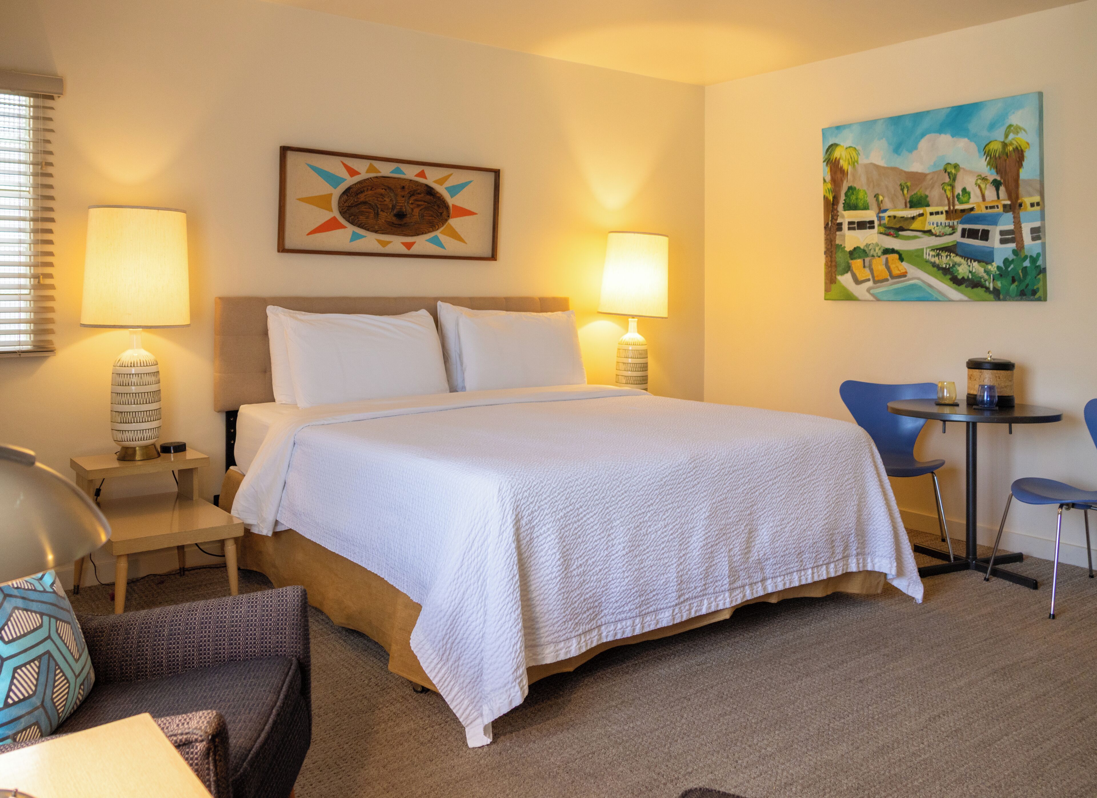 A hotel room with a bed, two bedside lamps, a small table with a lamp, a chair, and a painting on the wall.