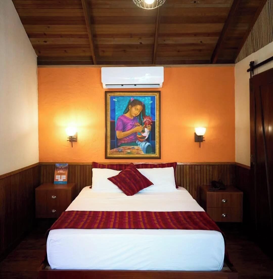 A hotel room with a bed, two bedside tables, a wall-mounted air conditioner, and two wall-mounted light fixtures.