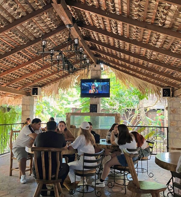 A group of people seated at tables under a wooden roof with a television screen mounted above. - Mayan Hills Resort (Copan Ruinas)