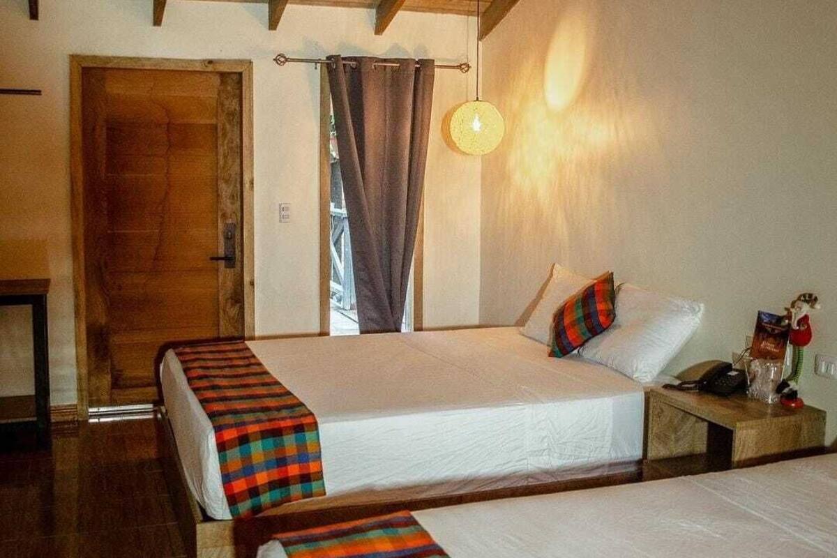 Quarto com duas camas, soalho em madeira, teto de madeira, uma porta e um candeeiro fixado na parede.
