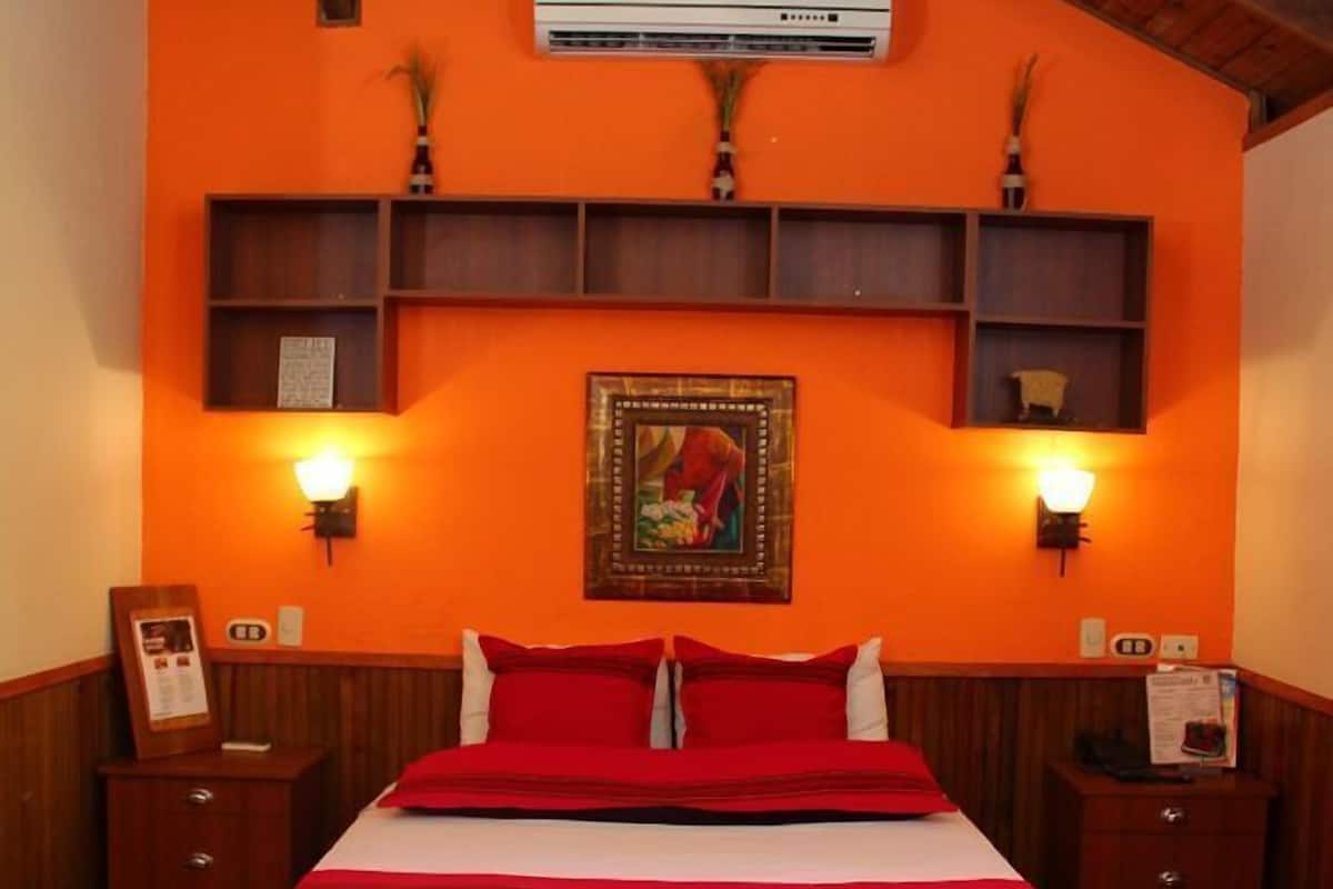 Quarto de hotel com teto de madeira, parede laranja, cama grande com roupa de cama vermelha, duas mesas de cabeceira e ar condicionado fixado na parede.