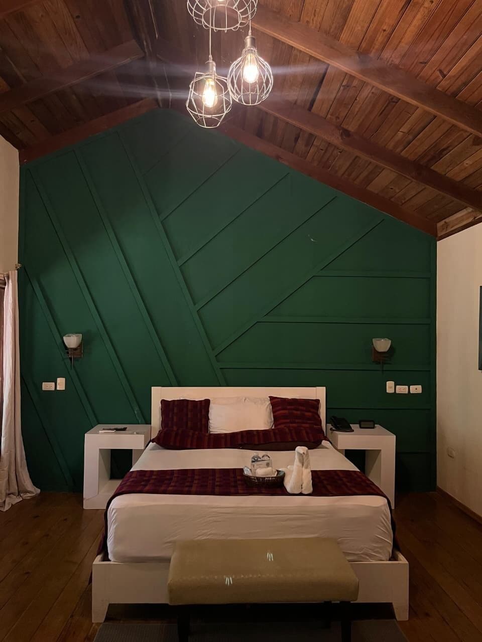 Un dormitorio con techo de madera, una pared de acento verde, cama blanca con ropa de cama roja y blanca, lámparas de noche y un banco al pie de la cama.