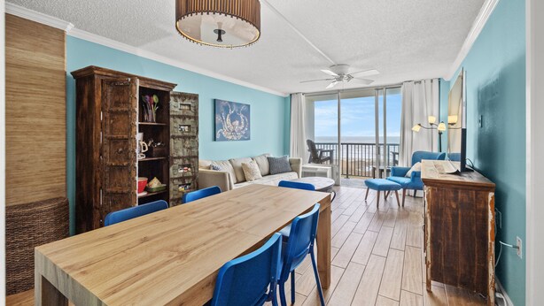 A dining area with a wooden table and blue chairs, a wooden cabinet with shelves, a ceiling fan, and a view of the outdoors through large windows. - East Island Oasis: On the Beach, NO Seawall Blvd, Mins 2 Everything, Pool, WIFI (Galveston)