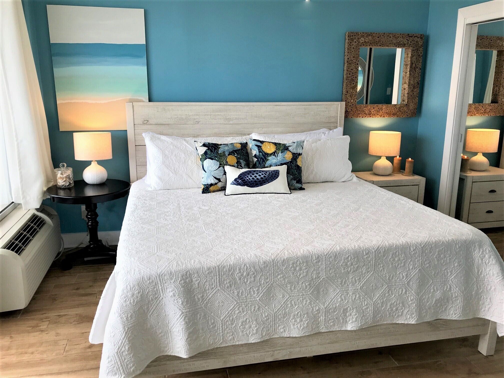 Una cama bien hecha con una colcha blanca y almohadas decorativas, una mesita de noche con una lámpara, y un espejo en la pared.