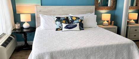 Una cama bien hecha con una colcha blanca y almohadas decorativas, una mesita de noche con una lámpara, y un espejo en la pared.