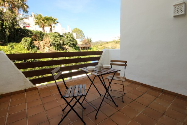 Un patio con un banco de madera y dos mesas, una con un cuenco blanco y otra con una jarrón blanco.
