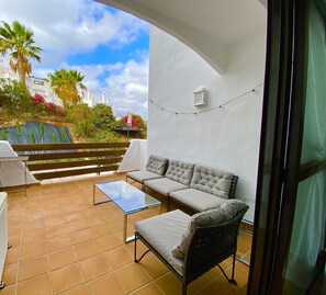 Ein Balkon mit Glastisch, gepolstertem Sofa und Korbstuhl. An der weißen Wand befinden sich ein Laternenlicht und Lichterketten.