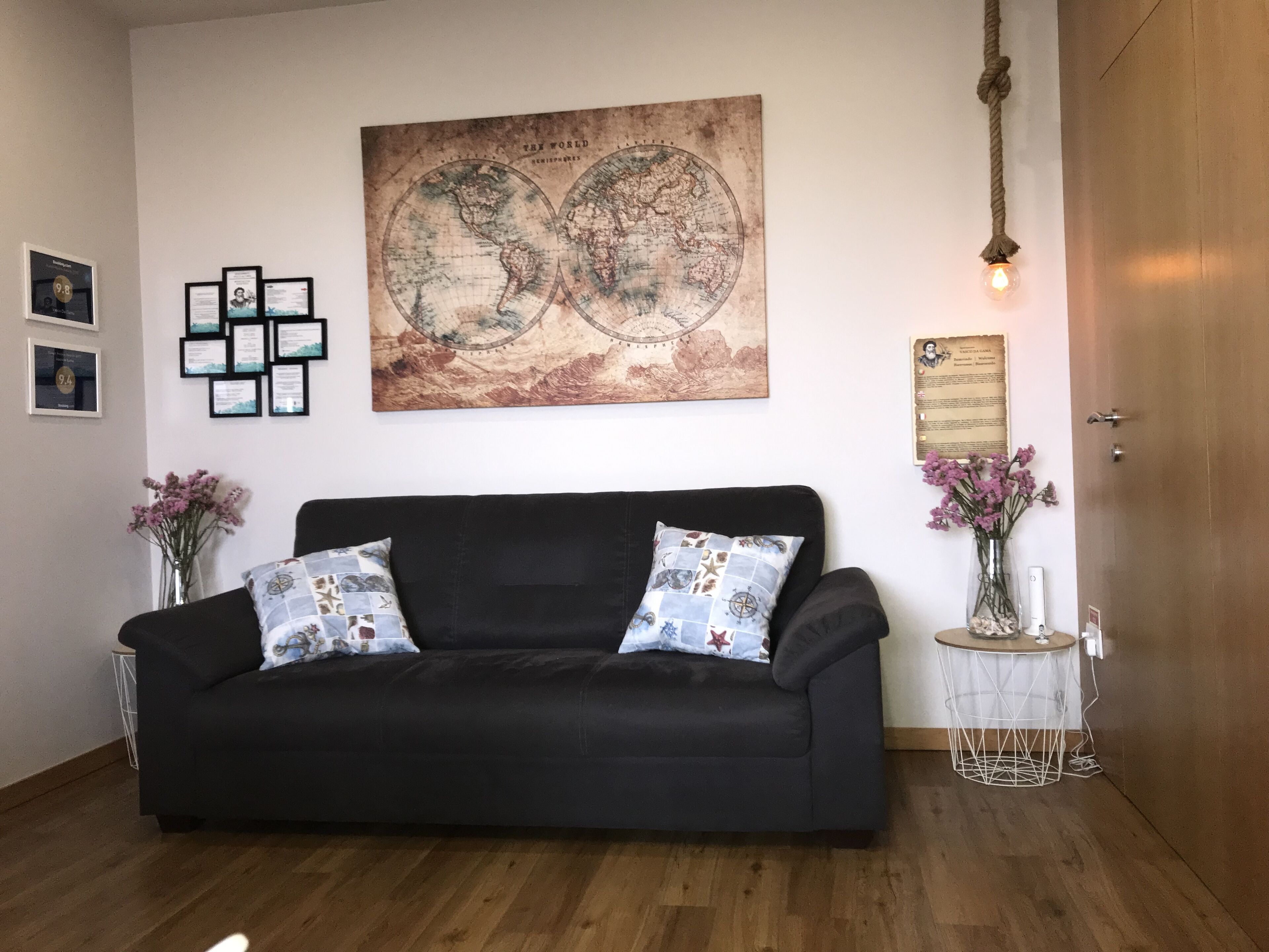 Un salón con un sofá negro, un mapa mundial en la pared, diplomas enmarcados, una mesita con un jarrón y una puerta de madera.
