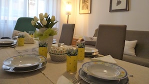 Zona pranzo con tavolo apparecchiato per quattro, piatti, bicchieri e un vaso con fiori. Si notano due sedie, un divano e una lampada sullo sfondo.