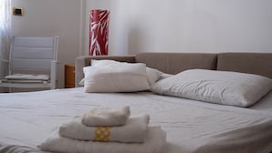 Un letto rifatto con lenzuola e cuscini bianchi, un asciugamano piegato, una sedia e un vaso con decorazioni rosse e bianche su una parete bianca.