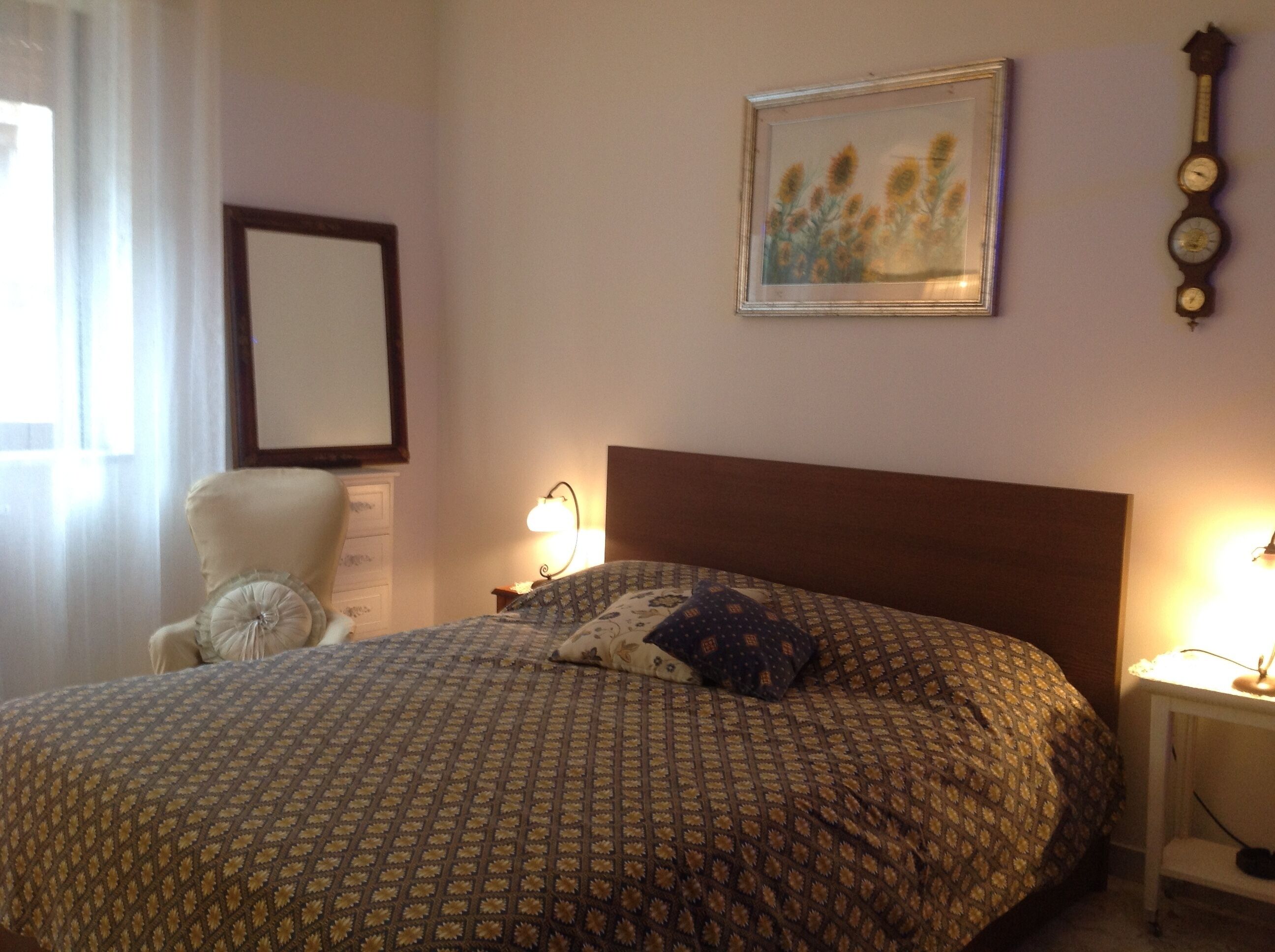 Ein Schlafzimmer mit einem Muster-Bettbezug, einem Sessel, einem Nachttisch mit Lampe, einem gerahmten Bild und einer Wanduhr.