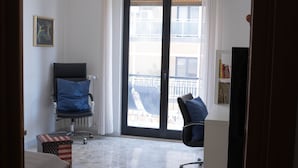 Una camera d'albergo con un'ampia porta-finestra scorrevole su un balcone, una scrivania con una sedia, un poggiapiedi con motivo a scacchi bianchi e rossi e un quadro appeso al muro.