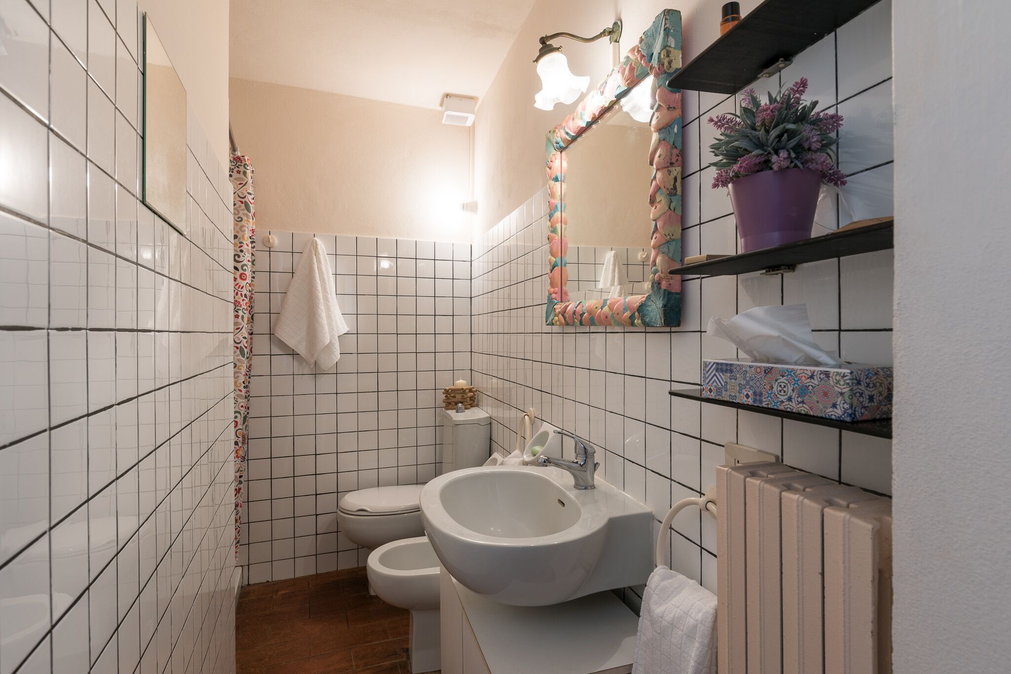 Un bagno con pareti piastrellate in bianco, un lavandino bianco, un water e una tenda da doccia. Sono presenti scaffali con articoli da toilette e una pianta in vaso.