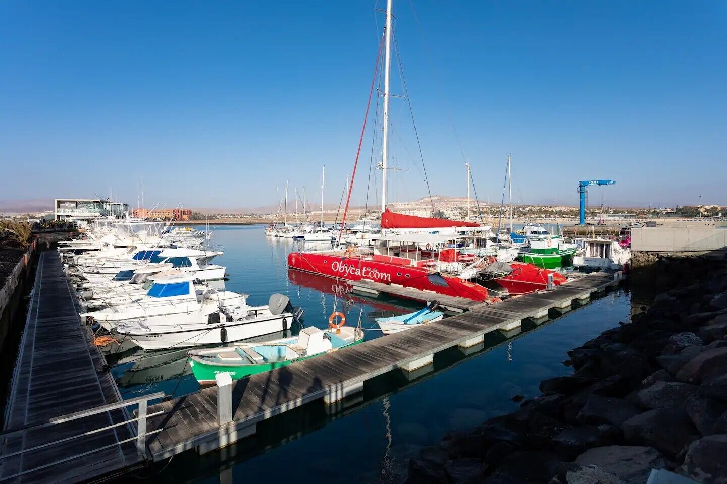 Un port de plaisance où sont amarrés différents types de bateaux, dont un voilier rouge sur lequel on peut lire le nom de domaine « Obycart.com ».