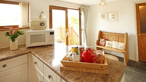 A kitchen with a microwave, a wooden bench with cushions, a tray with food items, and a window with curtains. - Beautiful 19th Century Seaside Cottage with hot tub and river views (Cemaes Bay, Anglesey)