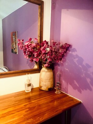 Ein Holzkonsolentisch mit einer Vase voller rosa Blumen, einem kleinen Glasbehälter und einem Spiegel, der eine violette Wand reflektiert.