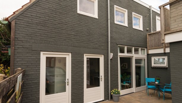 Une maison à deux étages, avec une façade de couleur gris foncé, des fenêtres à cadre blanc et un petit patio agrémenté de chaises bleues et d’une plante en pot.