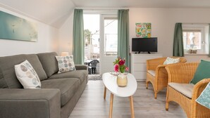 Un salon avec un canapé gris, des fauteuils en osier, une table basse blanche et un téléviseur posé sur un meuble.