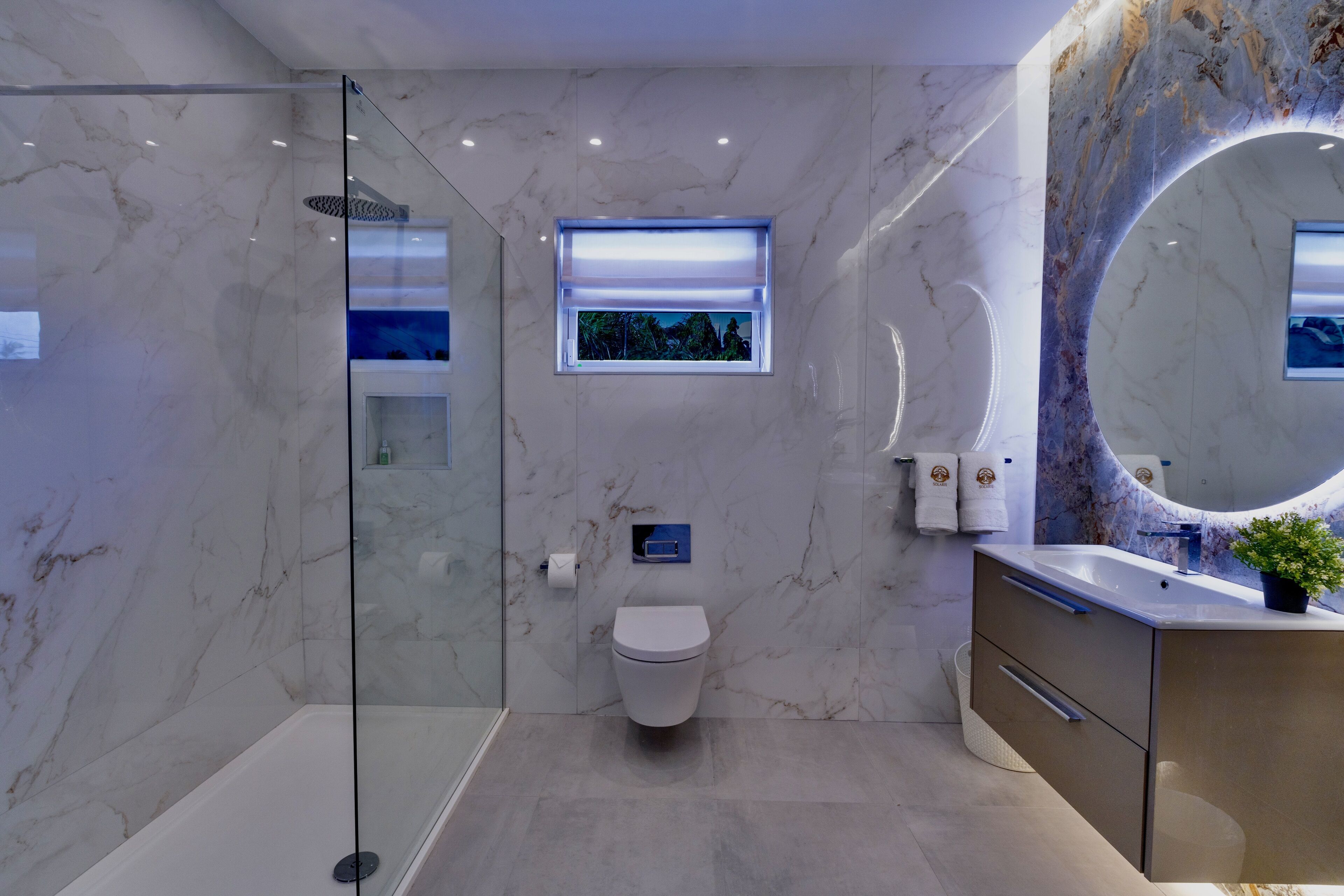 A modern bathroom with a glass-enclosed shower, a marble wall, a toilet, a sink with a large mirror, and a small window with a view.