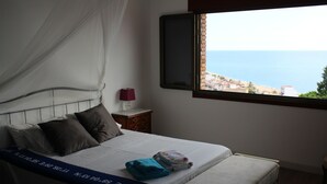 Ein Bett mit weißer Matratze, einer blauen Decke und Blick auf das Meer durch ein Fenster.