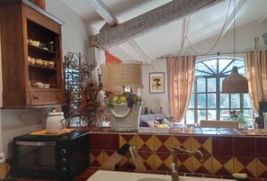 Een keuken met een houten kast, een magnetron, een spoelbak met een antieke kraan en een raam met gordijnen.