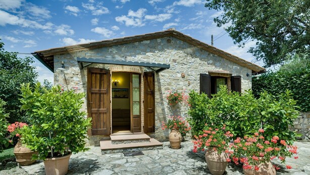 Una casa de piedra con puerta y contraventanas de madera, rodeada de plantas en macetas y un sendero de piedra.