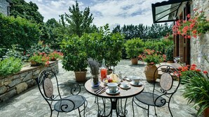Mesa redonda con sillas, rodeada de plantas y flores en macetas, sobre un patio de piedra con vista a árboles y un edificio.