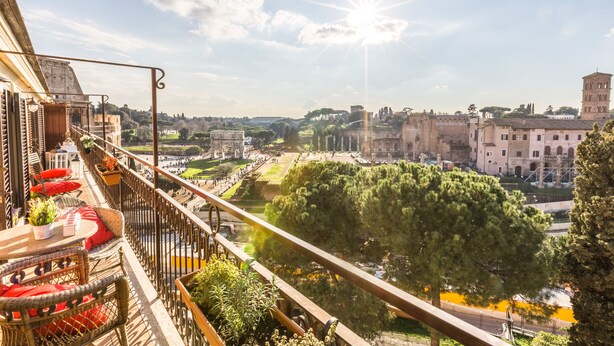 Un balcone con sedie in vimini e cuscini rossi, con vista su un centro storico caratterizzato da edifici antichi e un cielo sereno.