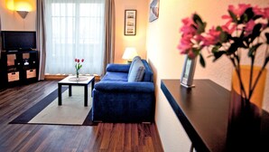 Uma sala de estar com um sofá azul, uma mesinha com um vaso de flores, uma luminária e uma televisão sobre um móvel.