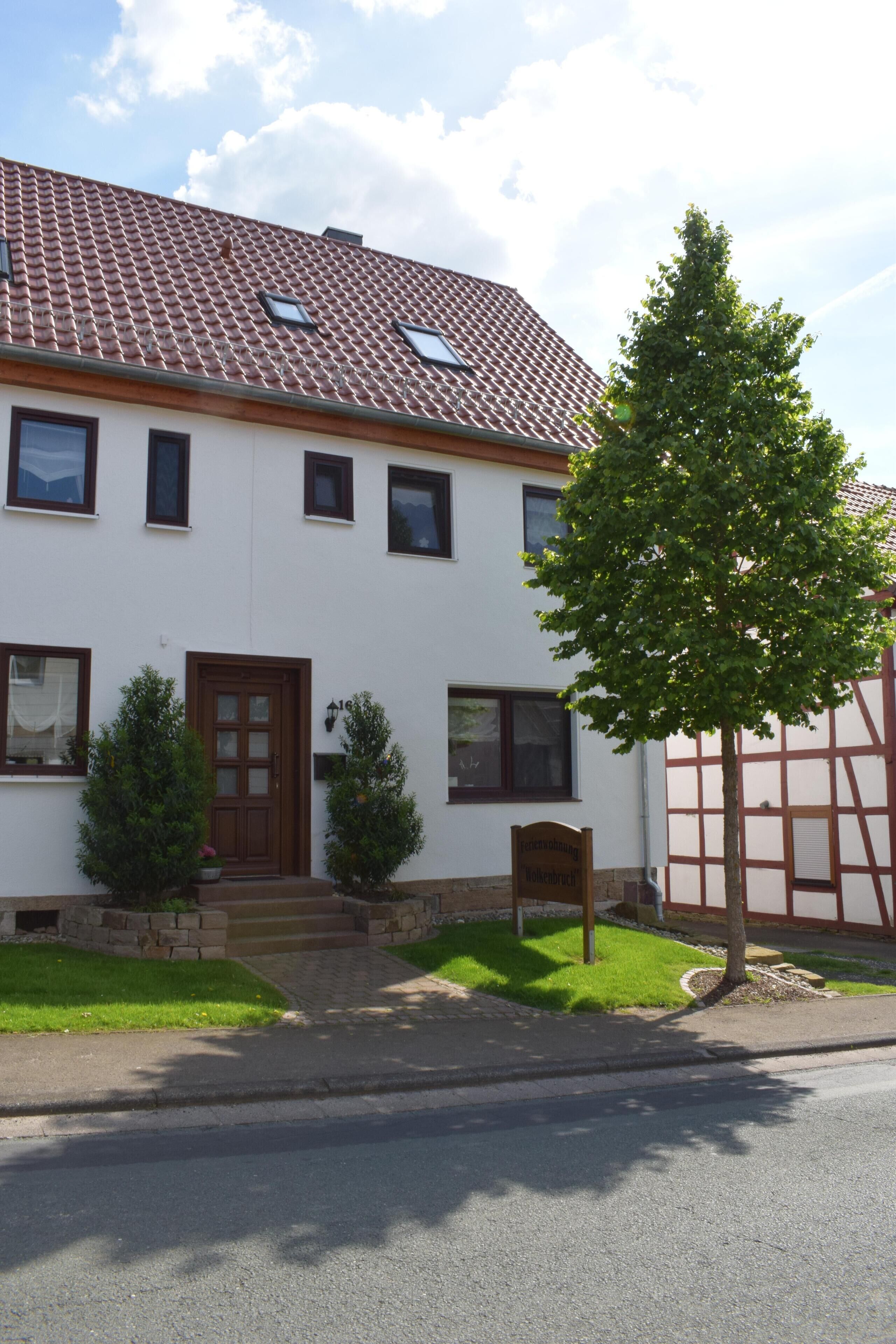 A two-story house with a tiled roof, a front door, and a small garden with a tree and shrubs.