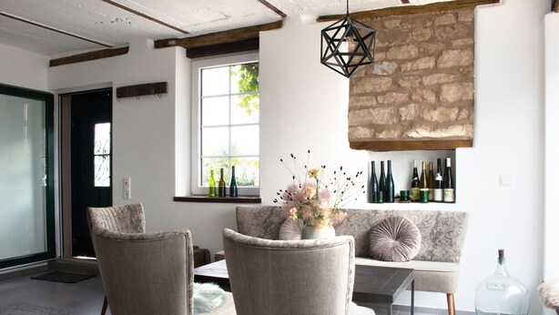 Ein modernes Wohnzimmer mit Steinwand, einer Couch, zwei Sesseln, einem Esstisch und einem Weinkühlschrank mit Flaschen.