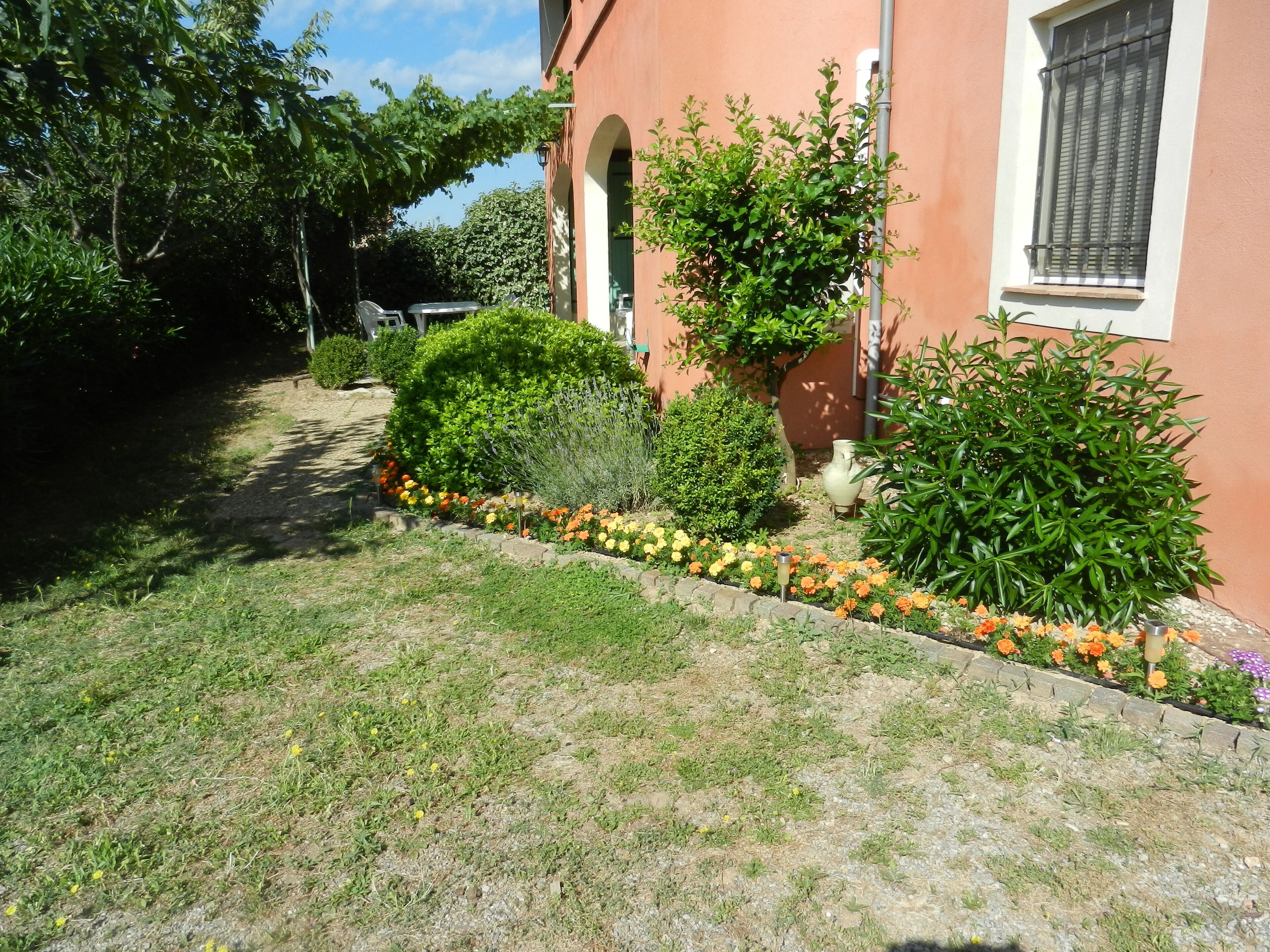 Un jardin bien entretenu, agrémenté de diverses plantes et fleurs, un chemin pavé et un bâtiment doté d’une entrée en arc de cercle.