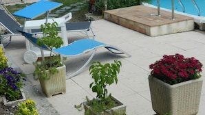 Zona junto a la piscina con sillas de descanso, plantas en macetas y solárium con baldosas.