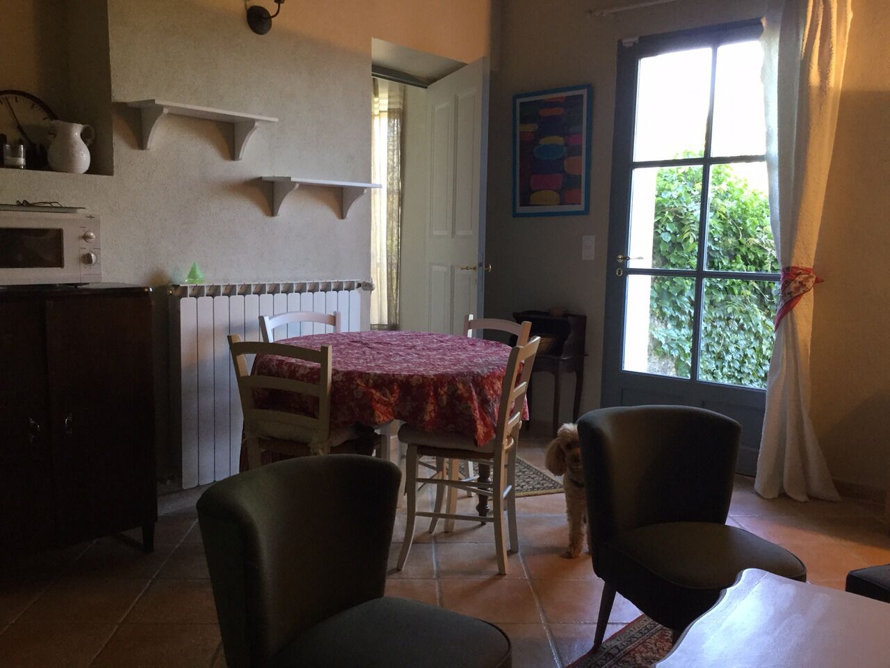 A dining area with a table covered in a red tablecloth, surrounded by chairs. There is a dog near the table, and a window with curtains is visible in the background.