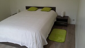 Een net opgemaakt bed met een witte deken en twee groene kussens, een nachtkastje met een lamp, en een groen vloerkleed.