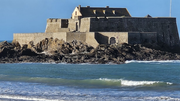 Een kustfortificatie met een opvallende toren en een wimpelstok, gelegen aan een rotsachtige kustlijn waar de golven op het strand breken.
