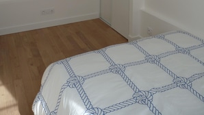 Een slaapkamer met een houten vloer, een bed met een deken met een wit en blauw patroon, een witte garderobe en een klein schilderij aan de muur.