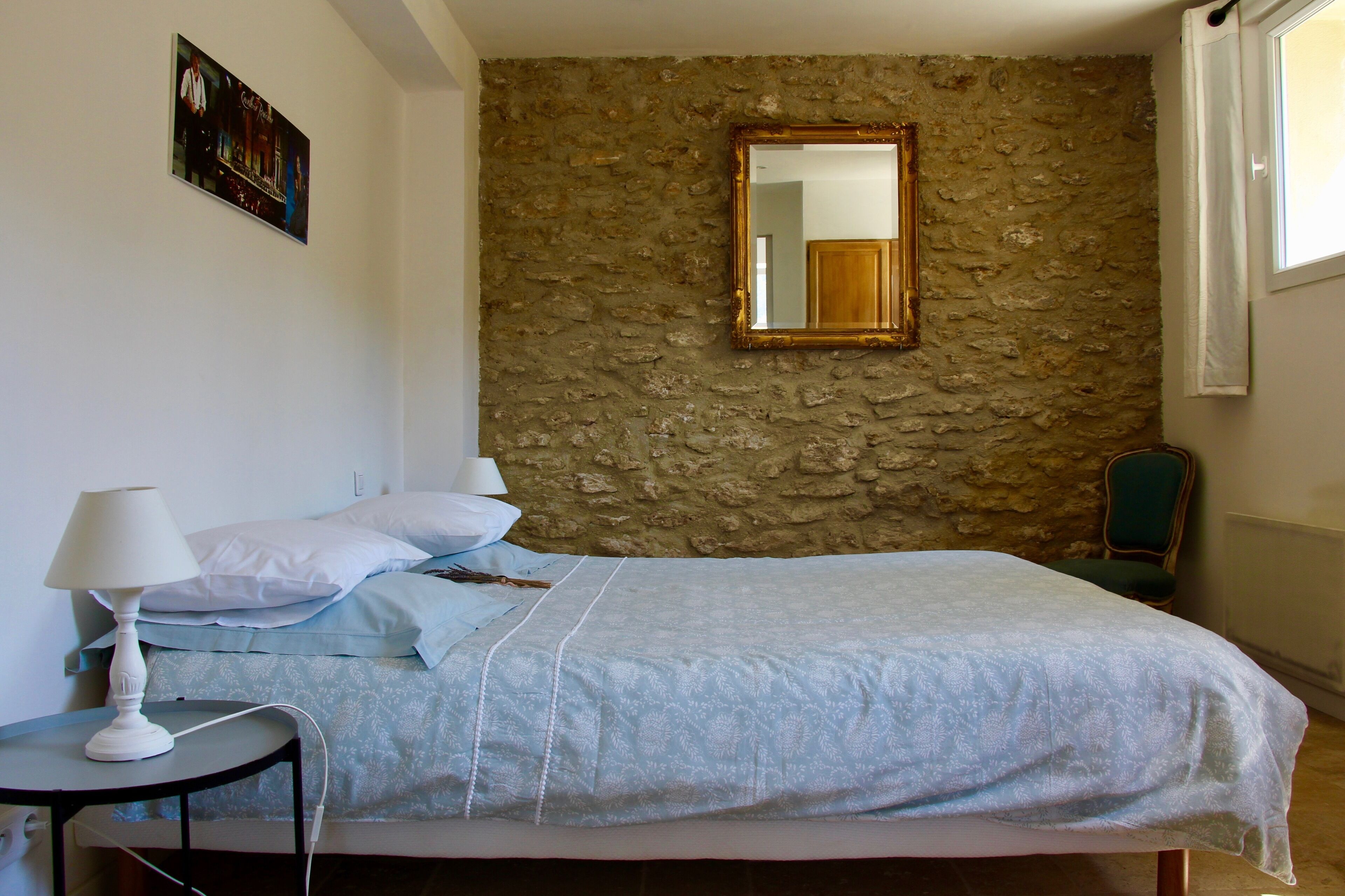 Une chambre avec un mur en pierre, un lit recouvert d’une courtepointe bleue, une table de chevet avec une lampe et un miroir.