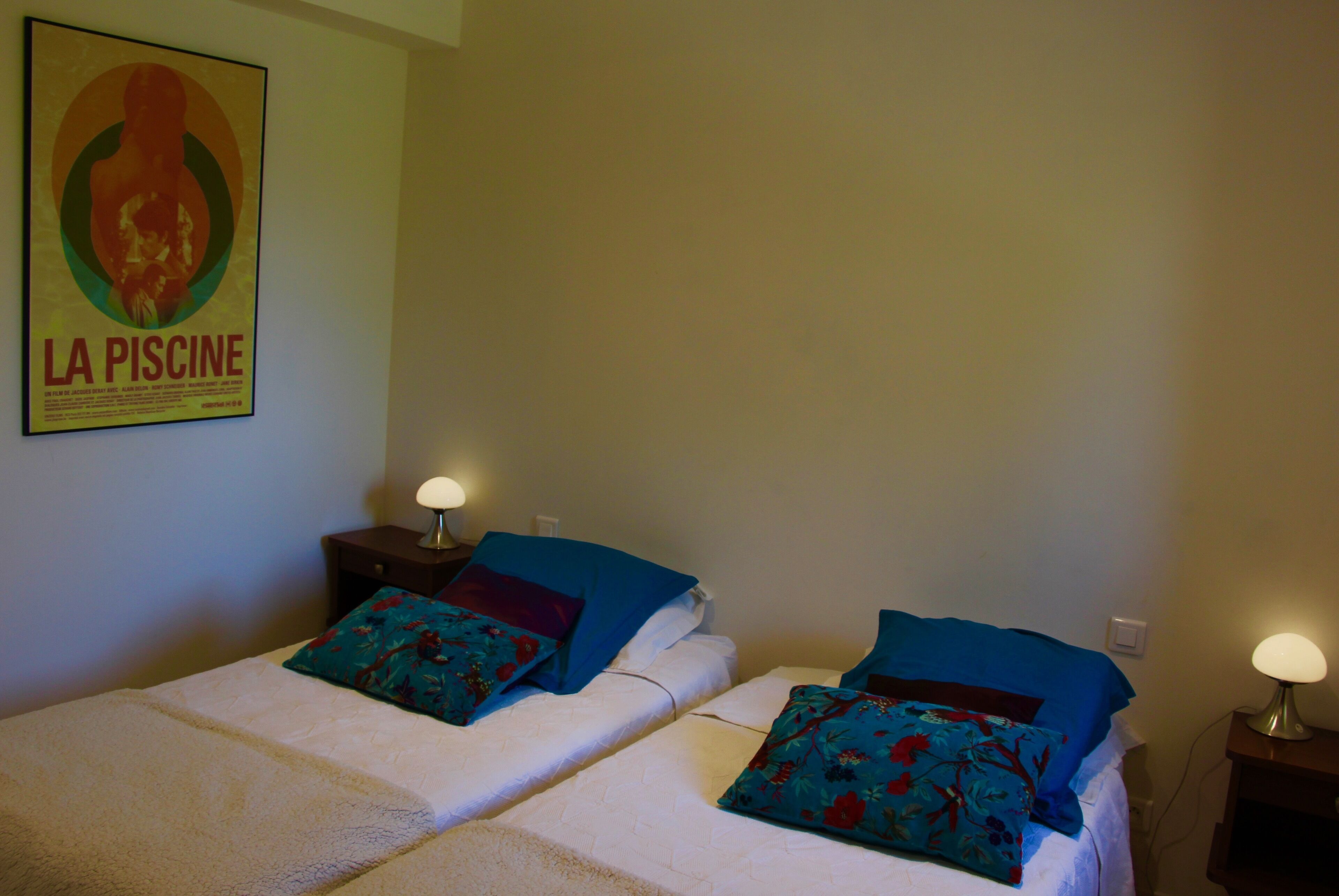 Een hotelkamer met twee bedden, elk met een bloemenkussen en een nachtkastje met een lamp. Een poster aan de muur luidt 