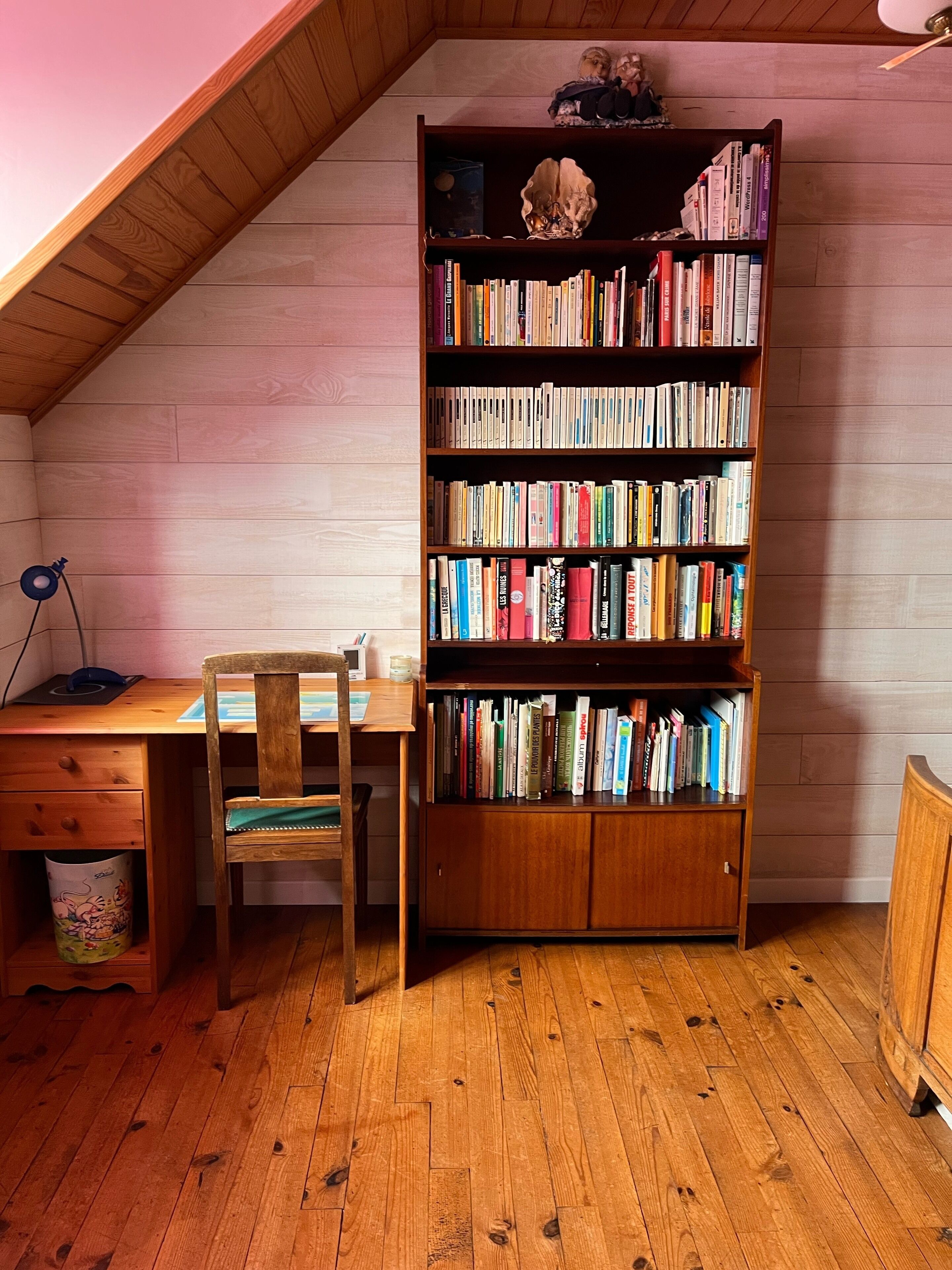 A room with a wooden desk, chair, and a tall bookshelf filled with books.