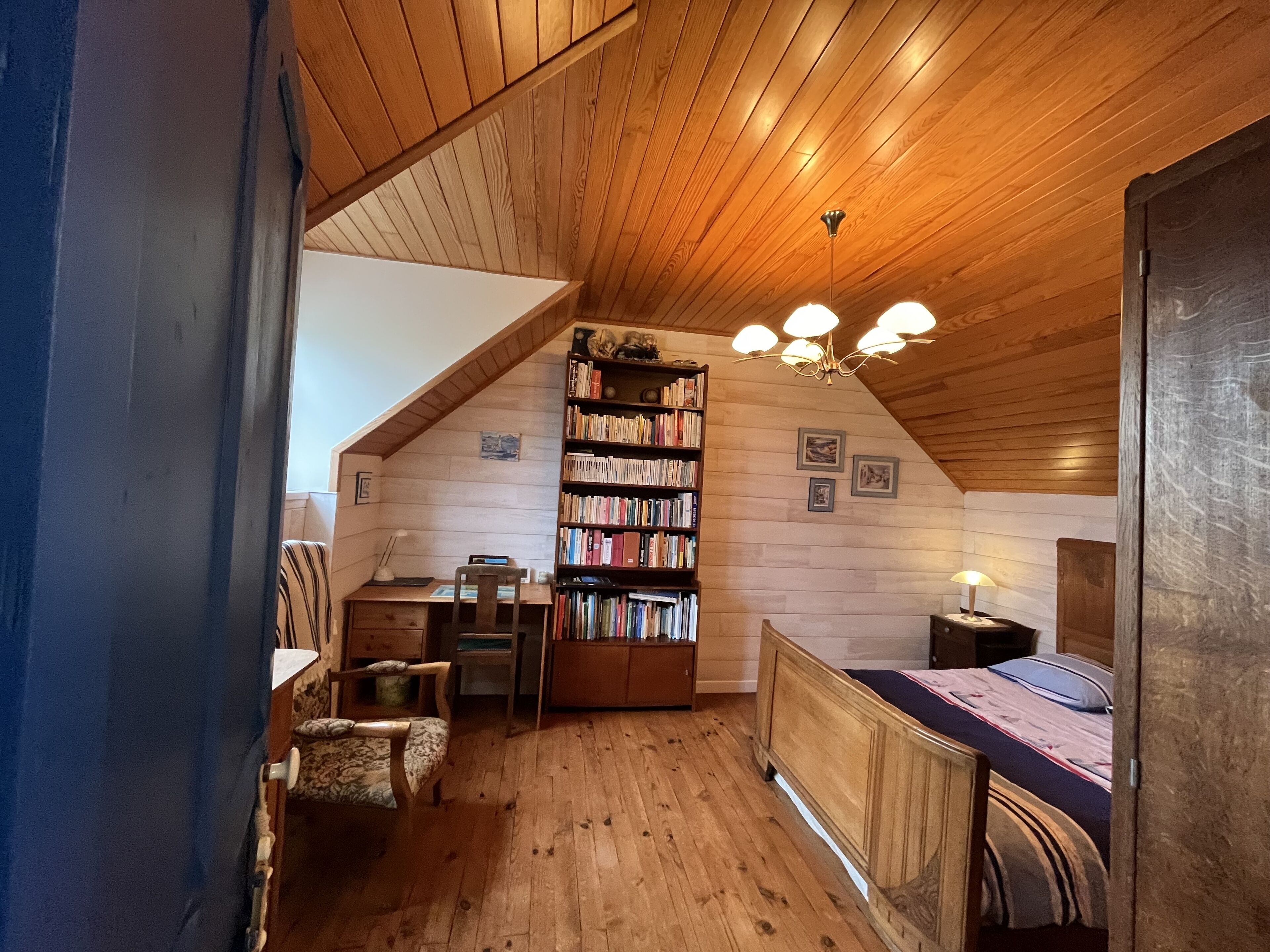 Een knus zolderkamer met houten wanden en plafond, een boekenkast, een bureau, een stoel en een bed.
